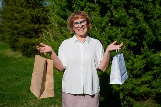 A happy elderly woman is standing in a park and holding paper bags. A retired shopaholic enjoys shopping. The concept of using eco friendly products to protect the environment