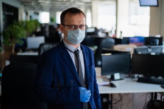 Serious man in a business suit and glasses in an empty office during quarantine. Male manager in a medical mask at the workplace. Social distance and workspace disinfection