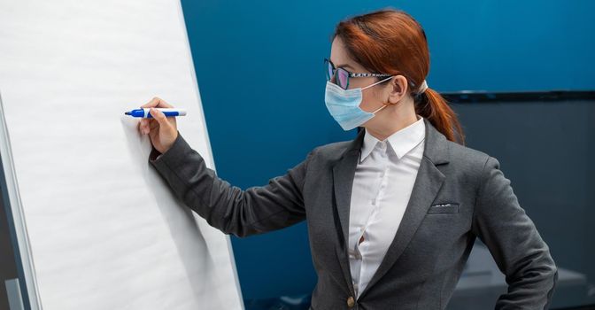 A woman in a medical mask leads a seminar in a conference hall. Business coach writes on a paper white board in the office. Female employee demonstrates presentation while maintaining social distance