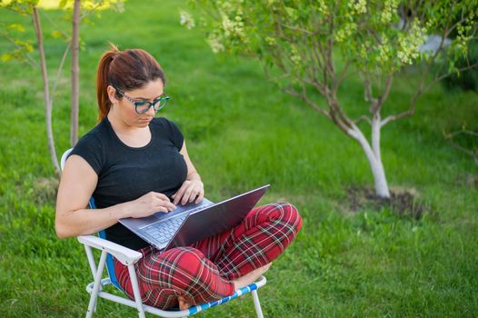 A woman with glasses sits in a folding chair with her legs crossed in a lotus position and typing on a laptop. Female freelancer remotely works in the garden of the cottage during quarantine