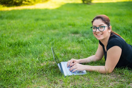 Happy red-haired girl in glasses lies on the lawn in the park and types on the laptop keyboard. Young caucasian female freelancer works remotely on a wireless computer outdoors
