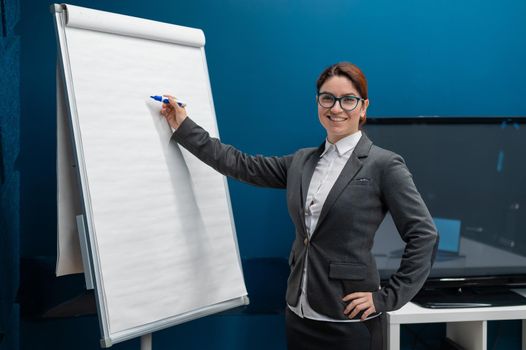 Friendly woman in a suit writes on a blank white board with a marker. Red-haired girl makes a presentation in the office. Beautiful female business coach at a conference.