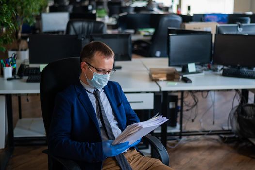 A man in a business suit and medical mask reads a paper report in an empty open space office. Social distance and isolation of employees. Urgent work during quarantine