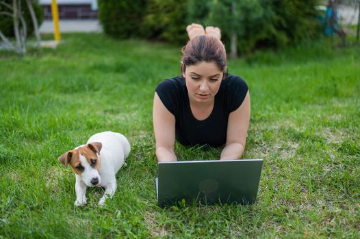 A red-haired woman lies on the green grass in a park with her own dog. The girl maintains a social distance and types on a laptop in the fresh air. A student is studying remotely on a computer