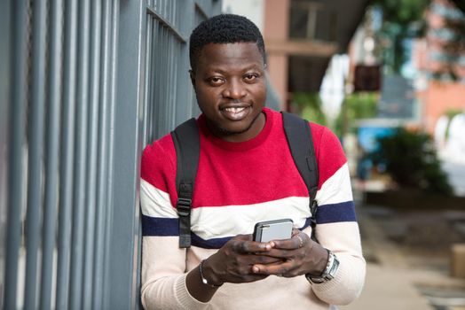 young student standing near grid with mobile phone backpack looking at camera smiling.
