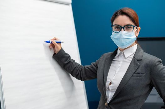 A woman in a medical mask leads a seminar in a conference hall. Business coach writes on a paper white board in the office. Female employee demonstrates presentation while maintaining social distance