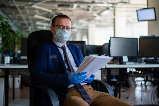 A man in a business suit and medical mask reads a paper report in an empty open space office. Social distance and isolation of employees. Urgent work during quarantine