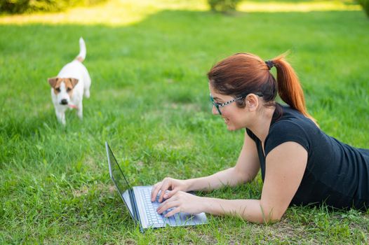 A woman with glasses lies on the green grass in the park with her own dog. A girl blogger maintains a social distance and maintains her blog outdoors. The student is studying remotely on a laptop