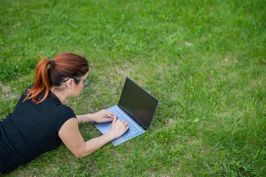 Young red-haired woman remotely works out lying on green grass. Freelancer girl prints on a laptop outdoors