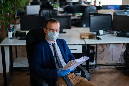 A man in a business suit and medical mask reads a paper report in an empty open space office. Social distance and isolation of employees. Urgent work during quarantine