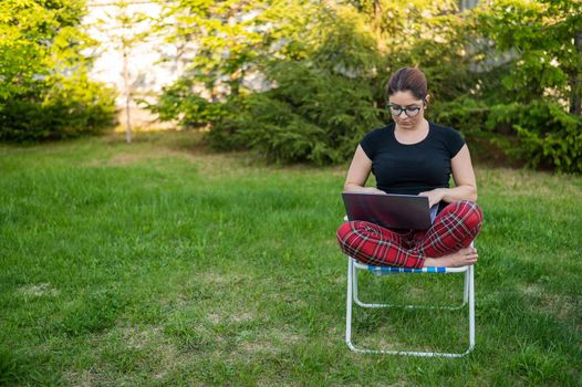 A woman with glasses sits on a folding chair and types on a laptop. Female freelancer works remotely in the garden of the cottage. Work in the open air during quarantine