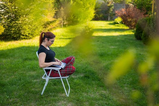 A woman with glasses sits in a folding chair with her legs crossed in a lotus position and typing on a laptop. Female freelancer remotely works in the garden of the cottage during quarantine