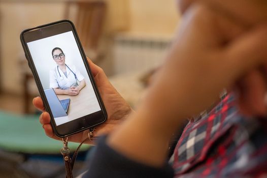 Rear view of an elderly faceless woman talking to a doctor via video chat. Portrait of a doctor on a mobile screen. Online medicine. Bed rest during quarantine