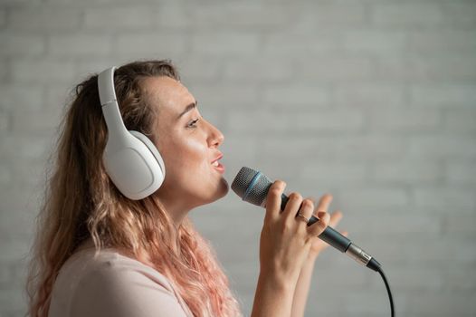 Close-up portrait of a caucasian woman with curly hair singing into a microphone. Beautiful sensual girl in white headphones sings a song in home karaoke