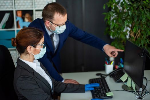 Colleagues in surgical masks in an open office space communicate at the work desk. A male top manager teaches a female intern how to work at a computer. The boss directs the subordinate