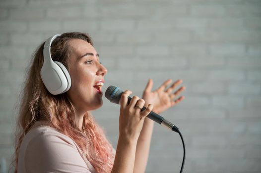 Close-up portrait of a caucasian woman with curly hair singing into a microphone. Beautiful emotional girl in white headphones sings a song in home karaoke and actively gestures against a brick wall