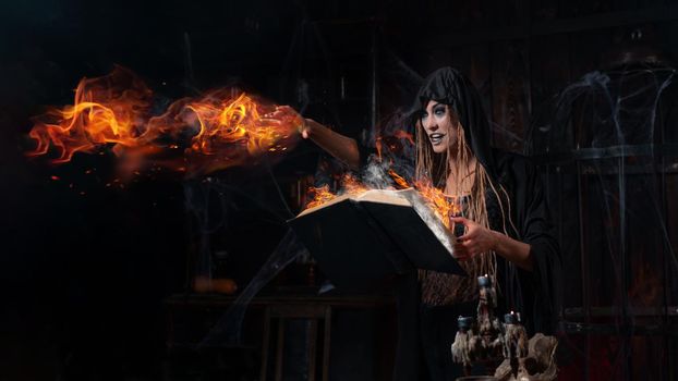 Halloween concept. Witch dressed black hood standing dark dungeon room use magic book for conjuring magic fire spell do fire ball. Female necromancer wizard gothic interior with cage, spider web