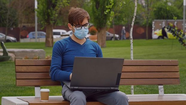 Young caucasian man in casual-wear sitting cross-legged on ground and working on laptop. Freelancer working outside typing on laptop computer. Red wall on the background.