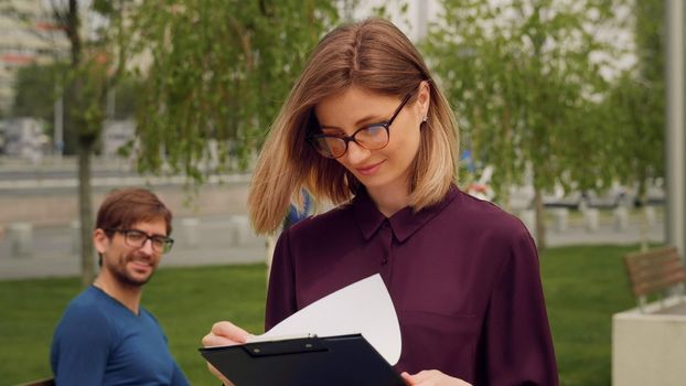 Confident Businesswoman walking out of Business building. Young blonde Woman greets friend sitting on a bench while the Walk. Success. Happy woman expression.