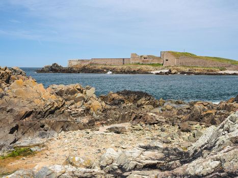 View across the rocky bay to Fort Grosnez from Fort Doyle under blue sky and white clouds, Alderney, Channel Islands