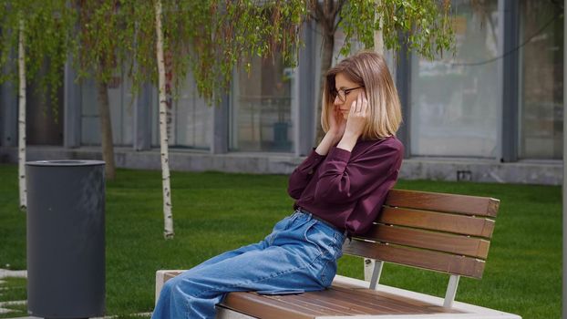 Woman suffering bad headache touching her temples outside sitting on a bench outdoor. Portrait of sad young Caucasian. Unhappy face.