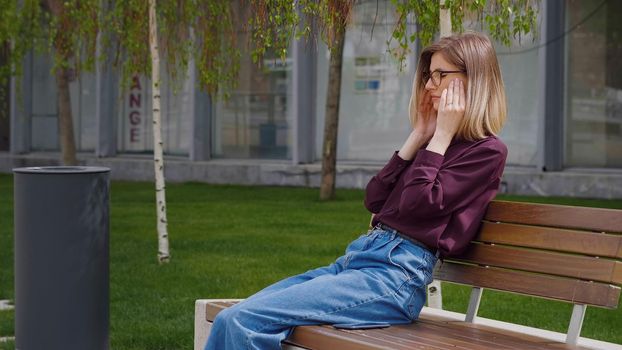 Woman suffering bad headache touching her temples outside sitting on a bench outdoor. Portrait of sad young Caucasian. Migraine. Unhappy face.