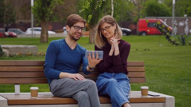 Young smiling couple conducting a video call on phone. Pretty Caucasian woman and man sitting outdoor beach coffee break. Phone video call conversation.