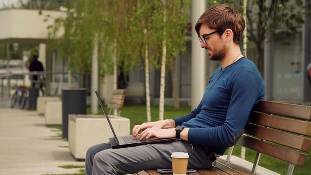 Young business man using laptop while sitting outdoor city park. Student studying on a bench. Entrepreneur working on laptop.