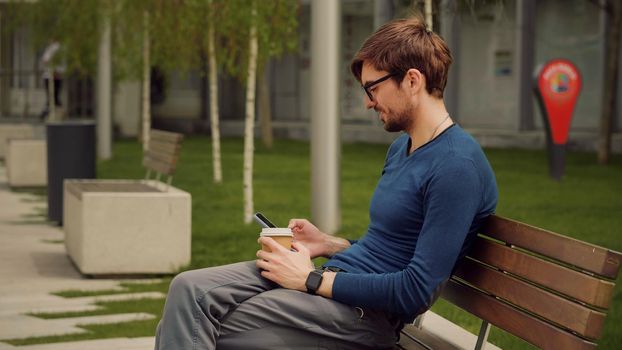 Outdoor scene of handsome young man browsing smartphone social network app while drink coffee. Student man relaxing in a park.