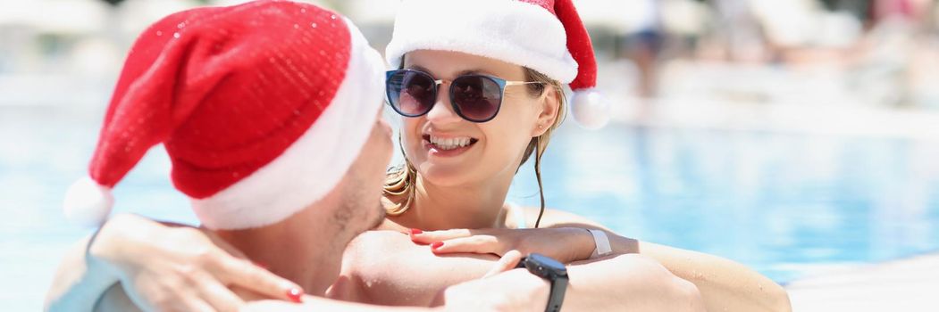 Portrait of woman hugging husband and smile to him wear santa claus hat. Couple resting on luxury resort, chill in swimming pool. New year, holiday concept
