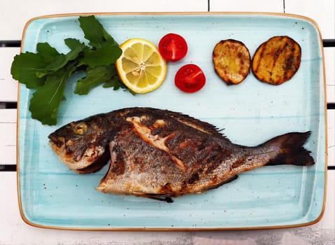 Fried fish with vegetables and herbs lies on light blue plate - tray, top view, close-up. Restaurant decoration of fish serving.