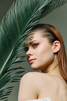 a sophisticated, elegant woman with evening makeup, stands with a green palm leaf, covering part of her face with it, looking away. Vertical photo without retouching. High quality photo