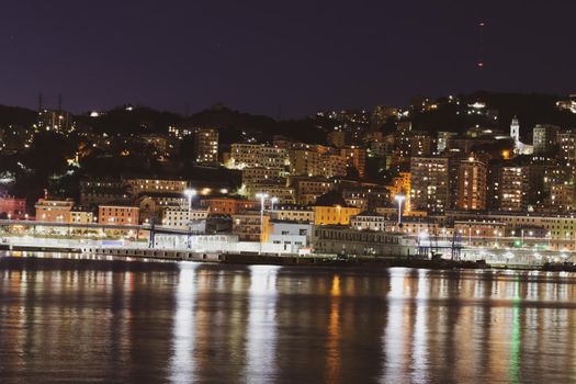 Genova, Italy - February 22, 2022: Evening view of Genoa (Genova) port, Italy with port cranes and industrial zone. Genoa, Italy. Lanterna lights by night. City illuminated behind the sea.