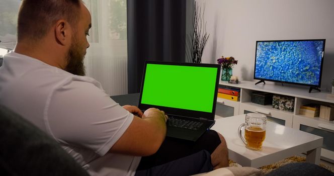 Man sitting on sofa Using Laptop with Greenscreen and Chroma Key. Mock Up with Tracking Markers and Alpha Matte. Caucasian Male relaxing in the evening on laptop and drink beer. Web search online.