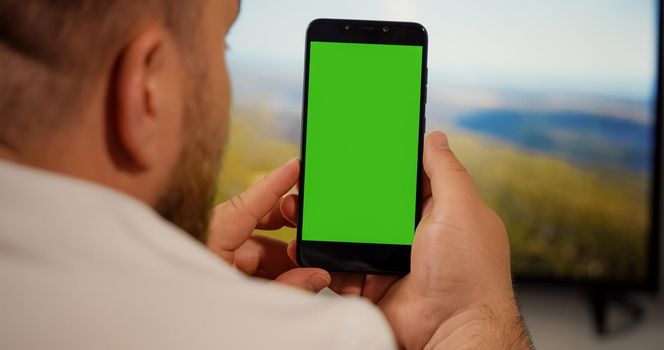 Man Watching on Smartphone Green Screen for Copy Space Chroma Key Mockup Tracking Markers and Alpha Matte while Sitting on living room couch. Close. TV video on background.