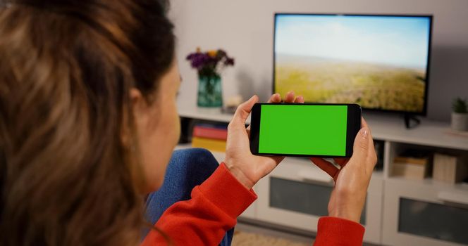 Phone Horizontal Green Screen for Copy Space Close up Chroma Key Mockup. Back over the shoulder view Girl watching video movie on couch Home. Hand phone holding technology users.