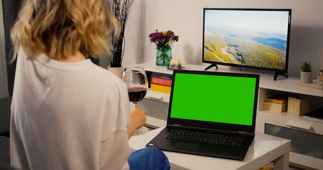 Over shoulder Laptop Green Screen for Copy Space Close up Chroma Key Mockup view. Happy woman has Video Call on with a Friend while Drinking a Glass of Wine. Relaxing Evening Conversation and wine.