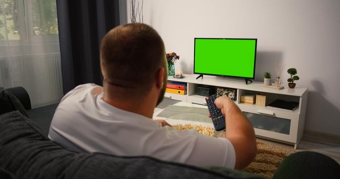 Man Watching Green Chroma Key Tv Screen and Changing Channels with Remote Control several times. Male sitting on the couch in the living room in the evening