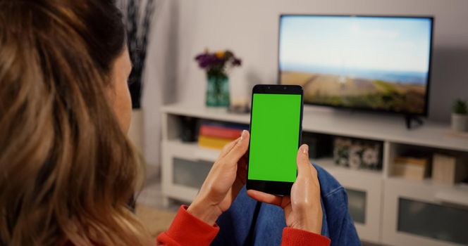 Woman at Home Sitting on a Couch using Smartphone with Green Mock-up Screen, Doing Swiping, Scrolling Gestures. Girl Using Mobile Phone, Internet Social Networks Browsing.