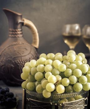 On the table in glass vase green grapes, near ceramic jug and two glasses guilt.