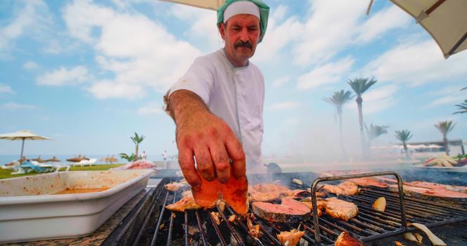 Tunisia, 2022: Chef cooking grilled salmon fillets for lunch, dinner for tourists of hotel. Beach resort. Outdoor grill plate with fish.
