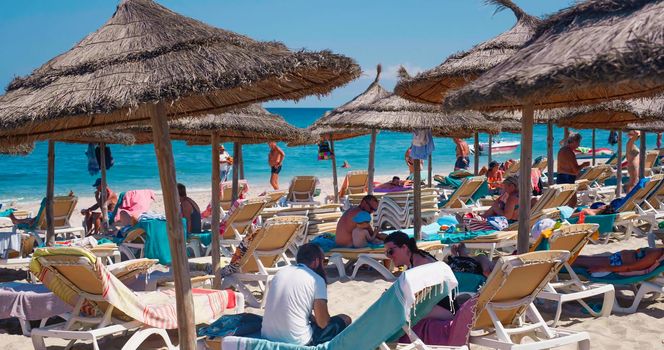 Tunisia, Africa, 2022: People relaxing on a summer day under sun umbrellas, in summer vacation. Chaise lounges with straw umbrellas on the beach. Luxury resort.