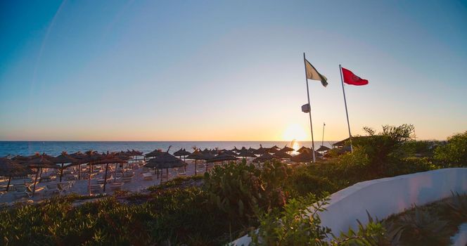 Sun loungers and umbrellas on the beach. Orange sky over sea. Sunset at deserted beach resort by sea in summer.