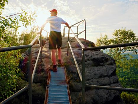 Tired hiker keep handrail on rocky view point.Sunny daybreak in rocky mountains. Hiker with red baseball cap, dark pants and white shirt. 