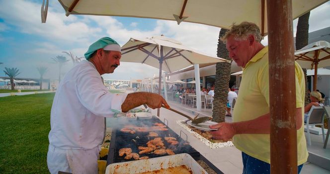 Tunisia, 2022: Chef cooking grilled salmon fillets for lunch, dinner for tourists of hotel. Beach resort. Outdoor grill plate with fish.
