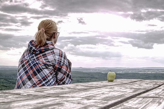 Young smiling blond girl trekker resting at wooden table with amazing view into landscape. Tired hiker sitting down and taking a break from hike. 