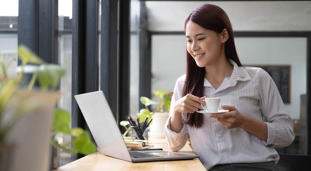 Beautiful young smiling Asian businesswoman working on laptop and drinking coffee, Asia businesswoman working document finance and calculator in her office..