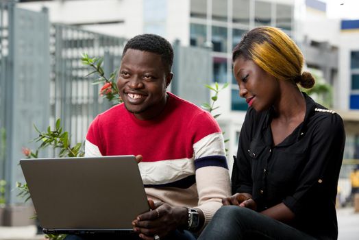 Two students sitting outside while working together on laptop on the side of the road outdoors in the summer.