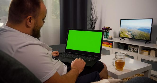 Man Using Laptop with Greenscreen and Chroma Key. Mock Up with Tracking Markers and Alpha Matte. Caucasian Happy Male has Video Call with a Friend Drinking Beer. Video conversation Friendship Concept.