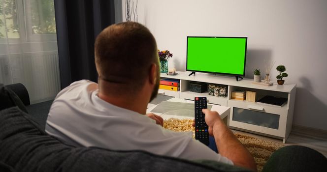 Back view Man sitting on the couch in the living room Watching TV with Green Screen and Changing Channels with Remote Control in his hand. Chroma Key TV in focus.